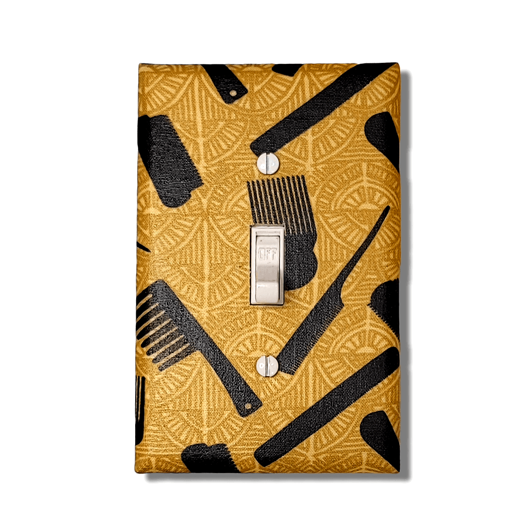 Handmade toggle light switch cover with rich gold fabric featuring intricate patterns of Black hair care tools including afro picks with fist handles, wide-tooth combs, brushes, and styling tools for Afrocentric cultural home décor