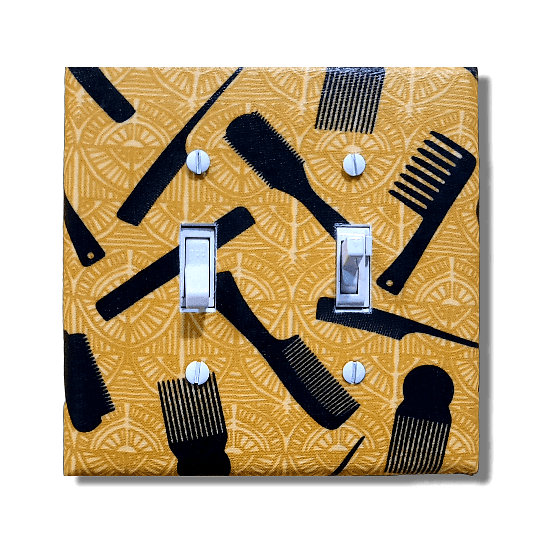 Handmade toggle light switch cover with rich gold fabric featuring intricate patterns of Black hair care tools including afro picks with fist handles, wide-tooth combs, brushes, and styling tools for Afrocentric cultural home décor